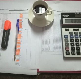 A workspace setting with financial documents, a black calculator displaying numbers, a cup of black coffee on a saucer, an orange highlighter, and a blue pen on a table.