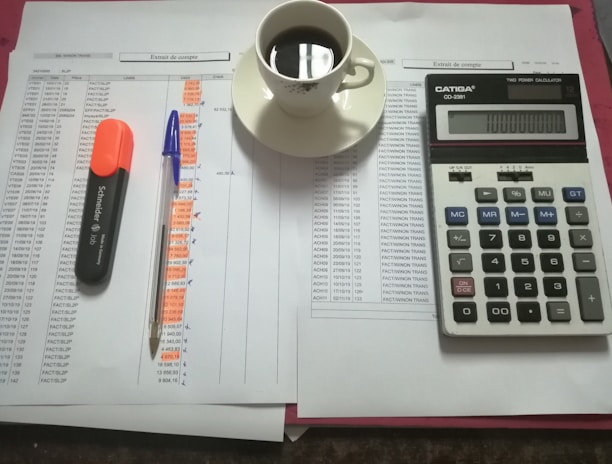 A workspace setting with financial documents, a black calculator displaying numbers, a cup of black coffee on a saucer, an orange highlighter, and a blue pen on a table.