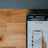 A close-up view of a smartphone displaying a financial app with a graph and numerical data, held by a hand. The screen shows the brand 'Wealthsimple'. Above it is part of a laptop keyboard with keys visible. The background consists of a wooden surface.