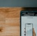 A close-up view of a smartphone displaying a financial app with a graph and numerical data, held by a hand. The screen shows the brand 'Wealthsimple'. Above it is part of a laptop keyboard with keys visible. The background consists of a wooden surface.