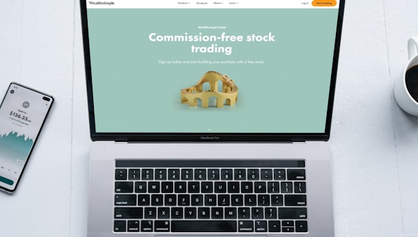 A laptop with a financial website open is centered on a white table. The screen displays a mint green background with text related to commission-free stock trading, accompanied by a gold object resembling a bridge. To the left, a smartphone displays a financial app with a graph and monetary figure. A cup of coffee sits on the right side, adding a casual work environment atmosphere.
