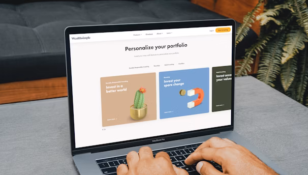 A person is typing on a MacBook Pro, viewing a website titled 'Personalize your portfolio.' The webpage displays three sections related to investing, each with different images and text. The laptop is on a grey table with a dark sofa and indoor plants in the background.